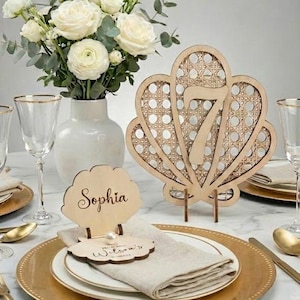 May include: A table setting with a floral centerpiece, gold-rimmed plates, and wooden seashell-shaped table number and place card holders. The table number is a seven, and the place card says "Sophia".