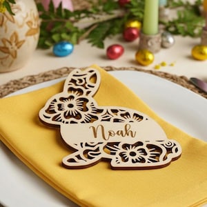 May include: A wooden bunny place card with the name "Noah" on a yellow napkin, set on a white plate. The bunny has a floral design. The table setting includes flowers, candles, and decorative Easter eggs.