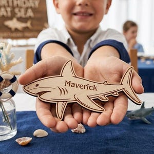 May include: A wooden shark-shaped name tag with the name "Maverick" engraved on it. The tag is held in a person's hands. The background includes a table setting with seashells and a "Birthday at Sea" sign.