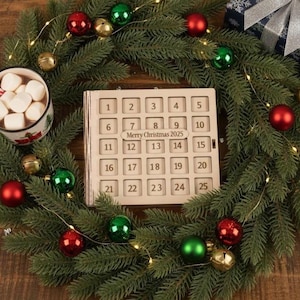 Puede incluir: Una corona navideña con adornos rojos, verdes y dorados rodea un calendario de adviento de madera. Una taza de chocolate caliente con malvaviscos y un bastón de caramelo está cerca. También hay una caja de regalo envuelta.