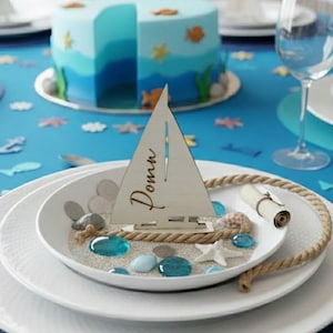 May include: A white ceramic plate centerpiece featuring a wooden sailboat cutout with the name Domn engraved on the sail. The plate is filled with sand, blue glass pebbles, small seashells, a white starfish, and a coiled rope accent piece.