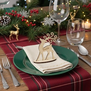 Puede incluir: Una mesa festiva con un árbol de Navidad al fondo. Una tarjeta de lugar de reno de madera se encuentra en un plato verde con una servilleta doblada. Copas de vino, cubiertos y velas completan la escena navideña.