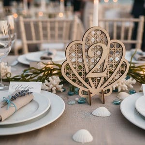 May include: Wooden table number 2 in a seashell design, with a woven pattern. The table setting includes white plates, folded napkins, and decorative elements like coral and seashells. A lit candle and wine glasses are also visible.