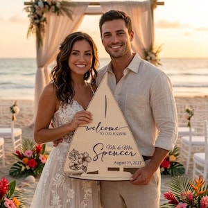 Puede incluir: Un letrero triangular de madera clara con la silueta de un velero, que presenta el texto grabado "welcome TO OUR FOREVER", "Mr. & Mrs. Spencer" y "August 23, 2027", junto con una ilustración floral detallada de flores de hibisco.