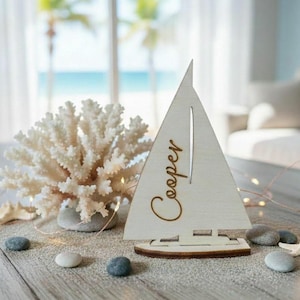 May include: A white wooden sailboat cutout featuring the name Cooper in cursive script, displayed alongside a white coral branch, several smooth gray pebbles, and a small white starfish on a light-colored wooden surface with string lights.