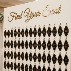 Może przedstawiać: Biała tablica z planem miejsc na wesele ze złotym napisem Find Your Seat u góry. Tablica zawiera rzędy czarnych, romboidalnych zawieszek wiszących na metalowych haczykach, z których każda ma nadrukowane imię gościa i numer stolika.