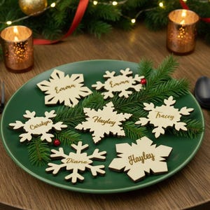 Op de afbeelding: Een groene bord met houten sneeuwvlok-naamkaarten, elk met een naam. Het bord is versierd met dennentakjes en is omringd door kaarsen, een vork en een lepel. Feestelijke vakantiedecoratie.