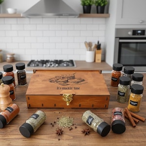Może przedstawiać: Drewniane pudełko na przyprawy ze złotym zapięciem i napisem "It's snackin' time" otoczone jest różnymi słoiczkami z przyprawami i drewnianym młynkiem do pieprzu. Słoiczki zawierają różne przyprawy, a niektóre są rozrzucone.