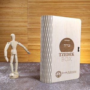 May include: A light-colored wooden Tzedek Box with the text "TZEDEK BOX" and a logo. The box is next to a wooden artist's mannequin and a small potted plant with purple flowers. The background is a wooden wall.