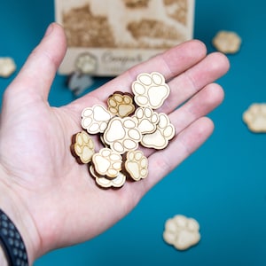 May include: A hand holding a pile of wooden paw print shaped game pieces. The pieces are light brown with white paw prints.