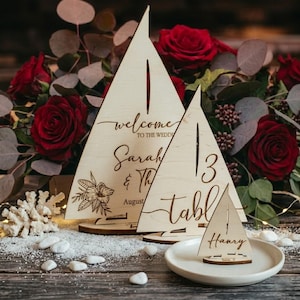 May include: Three light-colored wooden sailboat-shaped signs with laser-engraved text and floral designs. The largest sign reads welcome to the wedding Sarah & Thomas August, the medium sign reads table 13, and the smallest sign reads Henry.