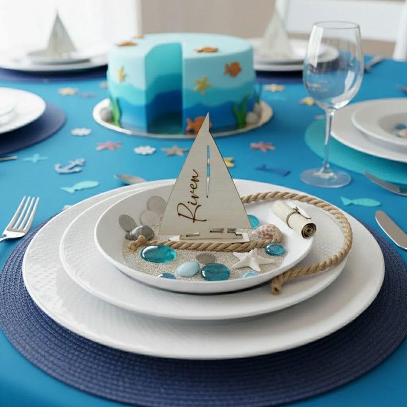 May include: A wooden sailboat place card with the name Riven engraved on the sail, displayed on a white plate filled with sand, blue glass pebbles, a small starfish, and a coiled rope, set on a table with a blue tablecloth and a cake.