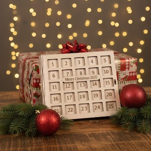 Könnte beinhalten: Ein hölzerner Weihnachtskalender mit nummerierten Feldern, einschließlich des Textes "Merry Christmas 2025". Der Kalender steht vor einem Geschenk mit roter Schleife, roten Kugeln und Grünzeug.