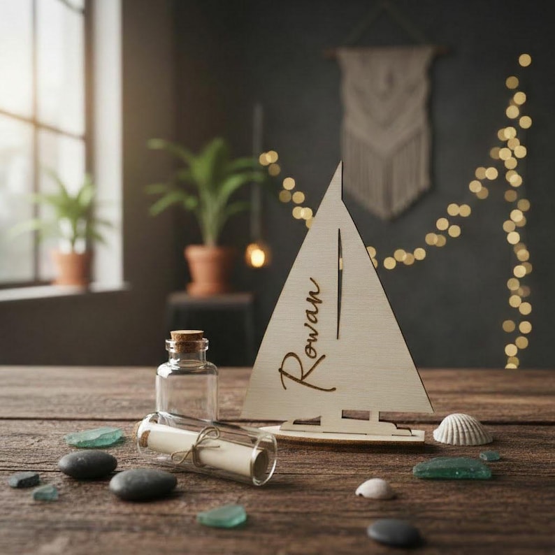 May include: A light-colored wooden sailboat cutout featuring the name Rowan in cursive script, accompanied by a small clear glass bottle containing a rolled paper scroll tied with twine, surrounded by several smooth dark and light sea stones.