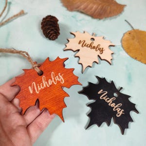Puede incluir: Tres etiquetas de madera con forma de hoja, cada una con el nombre "Nicholas" grabado. Una etiqueta es naranja, otra es de madera natural y otra es negra. Una piña y hojas de otoño están en el fondo. Las etiquetas están unidas a una cuerda.
