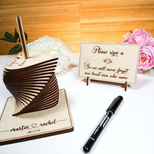 Op de afbeelding: Een houten gastenboekset met een stapel hartvormige stukjes op een vierkante voet gegraveerd met "martin & rachel 08.17.23" en een rechthoekig bordje gegraveerd met "Please sign a heart so we will never forget our loved ones who came".