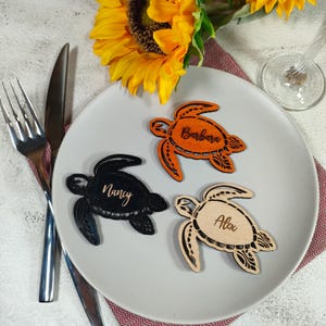 May include: Three wooden turtle-shaped place cards on a plate. One is black with "Nancy" in cursive, another is orange with "Barbara", and the last is natural wood with "Alex". A fork, knife, and sunflower are in the background.