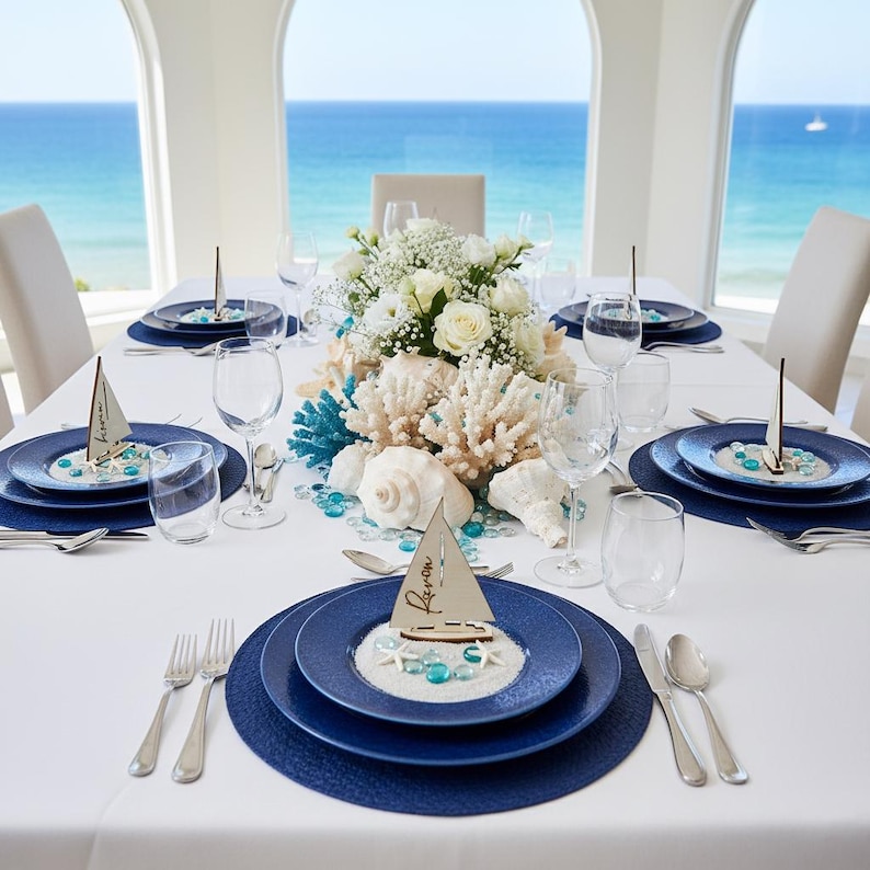 May include: A dining table setting featuring dark blue plates, clear glassware, and silver cutlery. Each place setting includes a small wooden sailboat cutout with the name "Raven" written on it, resting on a bed of white sand and blue glass beads.