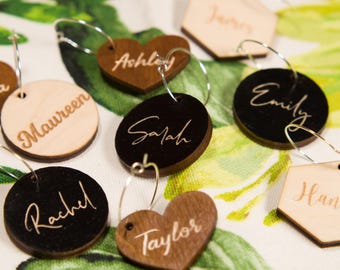 May include: A collection of wooden wine charms in various shapes, including circles, hearts, and hexagons. Each charm is engraved with a different name in a cursive font. The charms are in shades of brown and black, with silver wire rings for attachment.