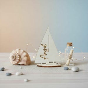 May include: A light-colored wooden sailboat cutout featuring the name Angel in cursive script, positioned next to a large spiral seashell and a small clear glass bottle containing a rolled paper scroll tied with twine, accented by thin fairy lights.