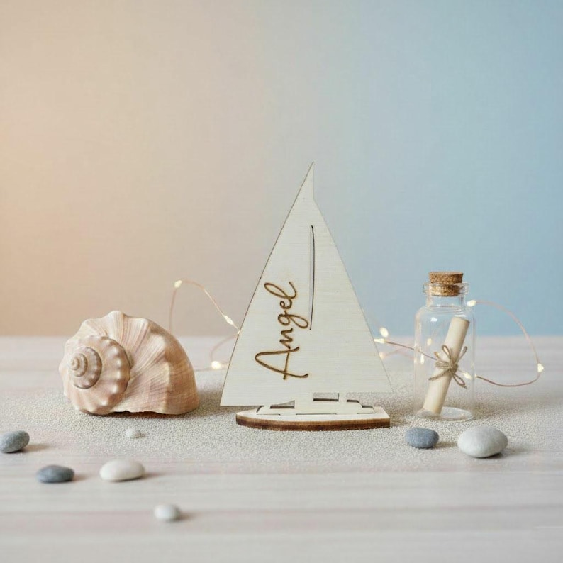 May include: A light-colored wooden sailboat cutout featuring the name Angel in cursive script, positioned next to a large spiral seashell and a small clear glass bottle containing a rolled paper scroll tied with twine, accented by thin fairy lights.