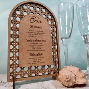 Peut inclure: Un panneau de menu de bar en bois avec une bordure décorative tressée. Le panneau énumère les recettes de cocktails : Old Fashioned, Cranberry Whiskey Sour et Kentucky Mule. Deux flûtes à champagne et un coquillage sont en arrière-plan.