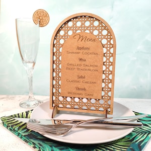 以下が含まれることがあります： 織り込まれたボーダーと茶色の背景にメニューが印刷された木製のメニューサイン。メニューには、前菜、メインコース、サラダ、デザートが含まれています。レモンスライスとカトラリーが入ったシャンパングラスが白いお皿の上にあります。