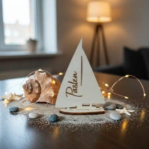 May include: A light-colored wooden sailboat ornament featuring the name Paslen engraved in a cursive script on the sail. The sailboat is surrounded by a large brown seashell, small white starfish, smooth gray pebbles, and thin copper fairy lights.