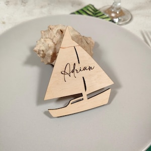 Puede incluir: Una tarjeta de lugar de madera en forma de velero, con el nombre "Adrian" grabado en la vela. La tarjeta de lugar es de color marrón claro y está colocada sobre un plato gris, junto a una concha. El diseño incluye detalles recortados para la estructura del barco.