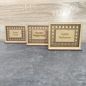 Puede incluir: Tres portatarjetas de comida de madera con texto. Las etiquetas dicen "Shrimp Cocktail", "Prosciutto Wrapped Melon" y "Stuffed Mushrooms". Cada etiqueta tiene un patrón tejido decorativo alrededor del texto.