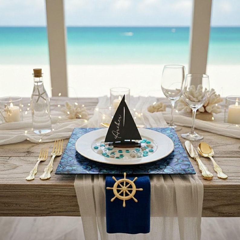May include: A table setting featuring a white plate with a black sailboat place card labeled Archer, resting on a blue iridescent placemat. A gold ship wheel napkin ring secures a blue napkin, accompanied by gold flatware and clear glassware.