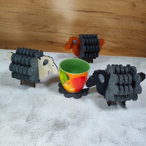 May include: A ceramic mug with a colorful apple design sits on a gray felt coaster, surrounded by three sheep-shaped felt and wood coasters. Two sheep are black, one is orange, and one is white.