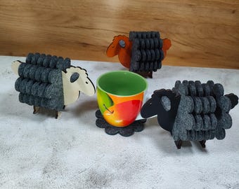 May include: A ceramic mug with an apple design sits on a grey felt coaster, surrounded by three sheep-shaped felt and wood coasters. Two sheep are black, one is orange, and one is white.