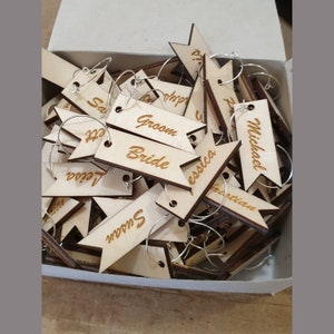 Puede incluir: Una caja llena de charms de madera para copas de vino con nombres grabados, incluyendo "Groom", "Bride", "Michael", "Jessica" y "Leila".