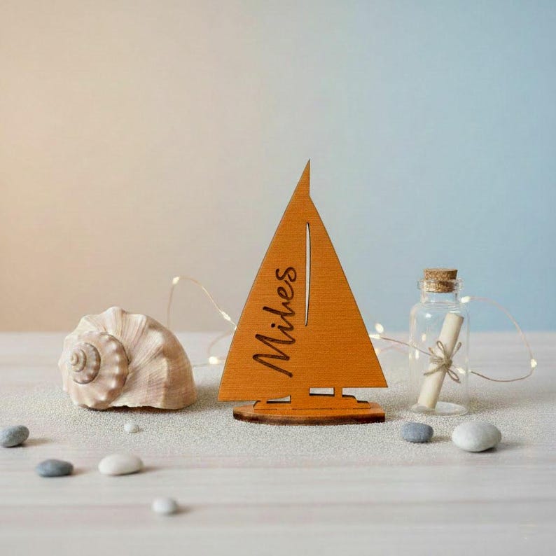 May include: A wooden sailboat cutout in a warm orange tone with the name Miles engraved on the sail. The sailboat is positioned next to a large spiral seashell and a small glass bottle containing a rolled paper scroll with a cork stopper.