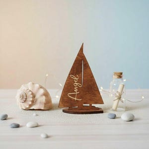 May include: A wooden sailboat cutout featuring the name Angel engraved in cursive script on the sail. The piece is accompanied by a large spiral seashell and a small clear glass bottle containing a rolled paper scroll tied with twine.