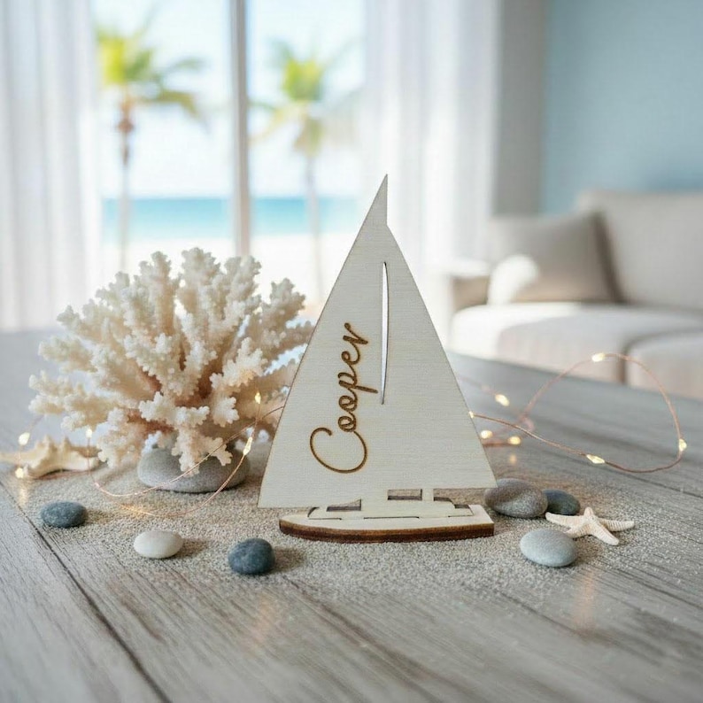 May include: A white wooden sailboat cutout featuring the name Cooper in cursive script, displayed alongside a white coral branch, several smooth gray pebbles, and a small white starfish on a light-colored wooden surface with string lights.