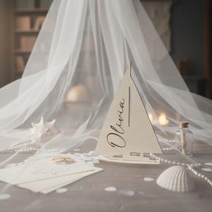 May include: A light-colored wooden sailboat cutout featuring the name Olivia in black script lettering. The sailboat is positioned alongside a white envelope, two gold rings, a white seashell, and a small glass bottle with a cork stopper.