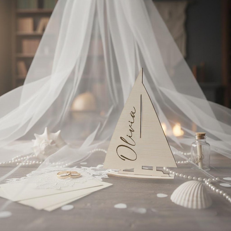 May include: A light-colored wooden sailboat cutout featuring the name Olivia in black script lettering. The sailboat is positioned alongside a white envelope, two gold rings, a white seashell, and a small glass bottle with a cork stopper.