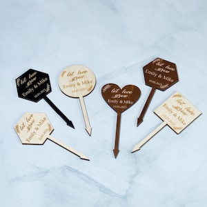 May include: A selection of wooden wedding signs in different shapes, including hexagon, circle, heart, and diamond. Each sign is engraved with the words "let love grow", the names "Emily & Mike", and the date "10.03.2023" or "10.03.2021".