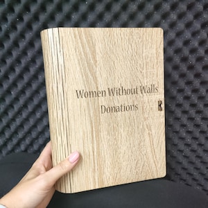 May include: A rectangular light brown wooden box with a vertical grain pattern. The front panel is engraved with the text "Women Without Walls" above the word "Donations". The box features a hinged side with visible vertical slats.