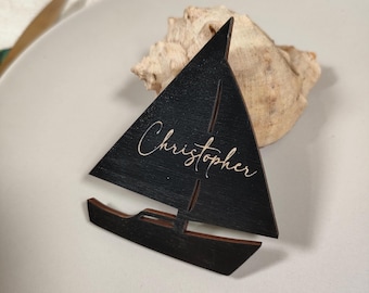 Puede incluir: Una tarjeta de lugar de madera negra con forma de velero con el nombre "Christopher" en fuente cursiva. La tarjeta de lugar está sobre un plato blanco con una concha en el fondo. El velero mide aproximadamente 10 cm de alto.