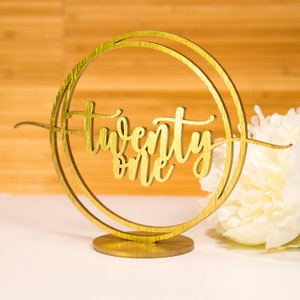 May include: A gold-colored wooden table decoration with the words "twenty one" in cursive script. The text is inside a double-ringed circle. The decoration is on a round base. A white flower is in the background.