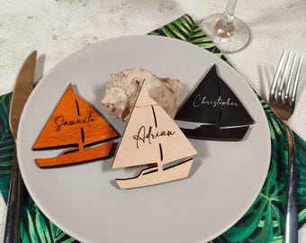 Puede incluir: Tres tarjetas de lugar de velero de madera en un plato. Una es naranja con "Samantha" escrita, otra es de madera natural con "Adrian", y la tercera es negra con "Christopher". Una concha marina se encuentra entre las tarjetas.
