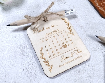 Puede incluir: Una tarjeta de madera con un calendario para guardar la fecha con un lápiz adjunto. El calendario es para junio de 2023 con un corazón marcando el 17. La tarjeta dice "Pencil us in... June 2023" y "Anna & Tom" con el texto "Formal invitation to follow".