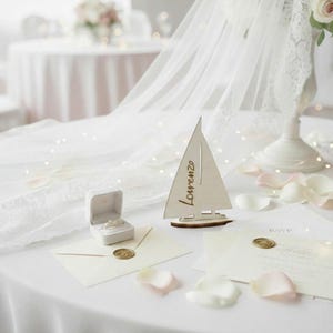 May include: A wooden sailboat-shaped table decoration featuring the name Lorenzo engraved in script, positioned next to a white ring box containing a ring, a cream-colored envelope with a gold wax seal, and a matching RSVP card on a white cloth.