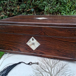 May include: A rectangular dark brown wooden box with a prominent wood grain pattern, featuring thin silver-toned inlay borders, a diamond-shaped mother-of-pearl keyhole escutcheon, and a separate metal key attached to a black tassel.