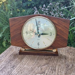 May include: A vintage Smiths clock with a wooden frame and a gold-tone clock face. The clock has a rectangular shape with a geometric design. The clock face features black numerals and hands. The clock is sitting on a wooden surface.
