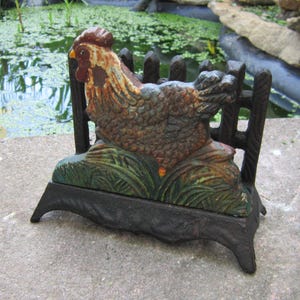 May include: A cast iron rooster-shaped garden ornament. The rooster is painted in shades of brown, blue, and orange, with a red comb. It is perched on a base with green grass and a black fence. The ornament is designed to hold something.
