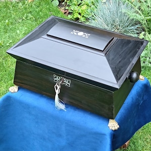 Peut inclure: Une boîte en bois sombre avec un couvercle à charnière et une petite serrure décorative. La boîte a une forme rectangulaire avec un dessus incliné et est ornée d'accents floraux. Elle repose sur de petits pieds dorés.